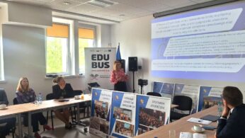 Kobieta stoi i przemawia do grupy siedzącej wokół stołu konferencyjnego w jasnym pomieszczeniu. Gestem wskazuje na ekran wyświetlający slajd prezentacji z polskim tekstem. Widoczne są banery i plakaty dotyczące edukacji i szkoleń zawodowych, a także okna i wystrój biura.
