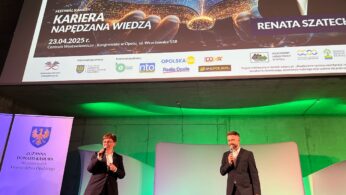 Dwie osoby w garniturach stoją na scenie i trzymają mikrofony. Za nimi znajduje się duży ekran wyświetlający reklamę festiwalu z cyfrową grafiką książki, szczegółami wydarzenia (23 kwietnia 2025 r., Opole) i zdjęciem kobiety. Niebieskie i zielone światła oświetlają scenę.