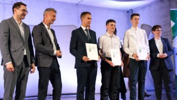 Sześć osób stoi na scenie. Trzech mężczyzn w garniturach stoi po lewej stronie, uśmiechając się. Trzech młodych mężczyzn w białych koszulach i ciemnych spodniach stoi po prawej stronie, trzymając certyfikaty i uśmiechając się. Tło jest oświetlone niebieskim i fioletowym światłem.