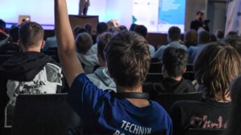 Grupa ludzi siedzi naprzeciwko sceny, na której przemawia prelegent. Na pierwszym planie osoba w niebieskiej koszuli podnosi rękę. Sceneria wygląda na konferencję lub seminarium z banerami i ekranem w tle.