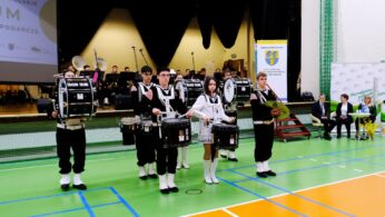 Orkiestra marszowa występuje na zielono-pomarańczowej podłodze sali gimnastycznej. Pięciu perkusistów i jeden cymbalista, wszyscy w czarno-białych strojach, stoją z przodu. Muzycy i urzędnicy siedzą i stoją na scenie i po prawej stronie, z transparentami i publicznością w tle.