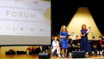 Dwie kobiety stoją na scenie, każda przy mikrofonie, zwracając się do publiczności. Za nimi widoczny jest siedzący zespół muzyków w mundurach. Po lewej stronie na dużym ekranie wyświetlany jest tytuł Kędzierzyńsko-Kozielskie Forum Edukacyjno-Gospodarcze.