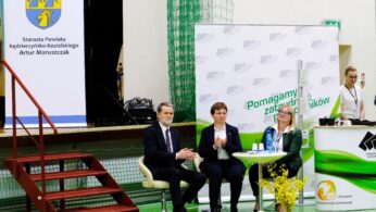 Trzy osoby w strojach biznesowych siedzą na scenie w siłowni. Dwie kobiety i mężczyzna siedzą obok małego stolika z papierami i butelkami wody. Widoczne są banery w języku polskim i żółta kompozycja kwiatowa; czwarta osoba stoi w pobliżu prawej krawędzi.