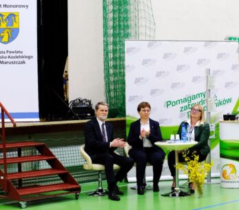 Trzy osoby w strojach biznesowych siedzą na scenie w siłowni. Dwie kobiety i mężczyzna siedzą obok małego stolika z papierami i butelkami wody. Widoczne są banery w języku polskim i żółta kompozycja kwiatowa; czwarta osoba stoi w pobliżu prawej krawędzi.