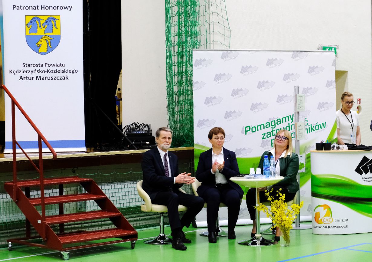Trzy osoby w strojach biznesowych siedzą na scenie w siłowni. Dwie kobiety i mężczyzna siedzą obok małego stolika z papierami i butelkami wody. Widoczne są banery w języku polskim i żółta kompozycja kwiatowa; czwarta osoba stoi w pobliżu prawej krawędzi.