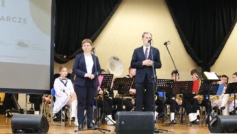 Dwóch mówców stoi na scenie z mikrofonami, ubranych w formalne stroje. Za nimi widoczny jest siedzący zespół młodych muzyków z instrumentami. Po lewej stronie znajduje się ekran projekcyjny z częściowym tekstem i logo. Tło sceny jest jasne z czarnymi zasłonami.