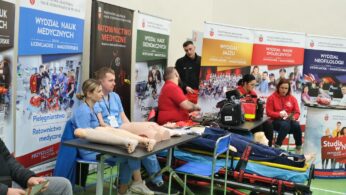 Grupa osób demonstruje opiekę medyczną w nagłych wypadkach przy użyciu manekinów CPR na targach edukacyjnych. Siedzą wśród sprzętu medycznego i kolorowych banerów różnych wydziałów akademickich. Obserwatorzy przyglądają się i robią notatki w jasno oświetlonym pomieszczeniu.