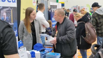 Kobieta w kraciastej marynarce stoi za stołem, rozmawiając z mężczyzną w czarnej kurtce na zatłoczonych targach pracy. Stół jest pokryty markowymi przedmiotami, broszurami, długopisami i niebieską czapką. W tle widoczne są inne osoby i stoiska.