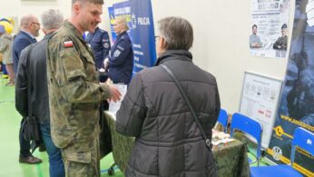 Żołnierz w mundurze kamuflażowym rozmawia ze starszą kobietą w czarnym płaszczu podczas wydarzenia wewnątrz budynku. Stoją w pobliżu stołu z materiałami informacyjnymi i plakatami w języku polskim. Inni uczestnicy i niebieskie krzesła są widoczne w tle na zielonej podłodze.