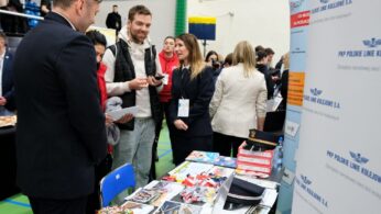 Ludzie stoją i rozmawiają na stoisku targów kariery PKP Polskie Linie Kolejowe S.A. Na stole rozłożone są broszury informacyjne, słodycze i drobne artykuły promocyjne. W tle widoczny jest tłum i sala gimnastyczna.