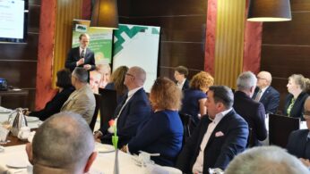 Mężczyzna w garniturze stoi i przemawia do siedzącej publiczności w nowoczesnej sali konferencyjnej. Ludzie, głównie w strojach biznesowych, patrzą na niego uważnie. Za mówcą znajdują się banery, a na stołach białe obrusy, filiżanki i pojedynczy tulipan w wazonie.