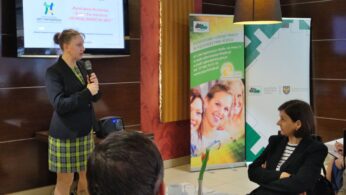 Kobieta stoi i mówi do mikrofonu w sali konferencyjnej. Stoi twarzą do siedzących uczestników. Za nią znajdują się banery z uśmiechniętymi twarzami i polskim tekstem oraz ekran wyświetlający informacje. Stoły są zastawione białymi obrusami, kubkami i talerzami.
