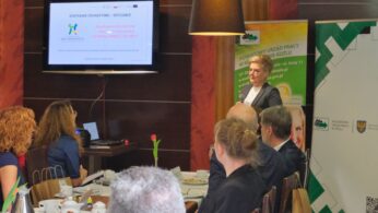 Kobieta stoi i prezentuje grupie siedzącej przy stolikach z talerzami śniadaniowymi. Ekran za nią wyświetla polską prezentację. Uczestnicy uważnie słuchają. Otoczenie to nowoczesny pokój z banerami i plakatami informacyjnymi w języku polskim na ścianach.
