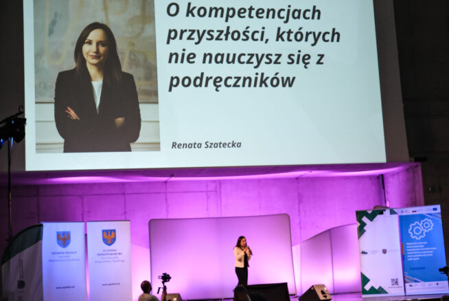 Kobieta stoi na scenie i mówi do mikrofonu. Za nią znajduje się duży ekran pokazujący jej zdjęcie, nazwisko (Renata Szatecka) i polski tekst o przyszłych umiejętnościach, których nie uczą w podręcznikach. Banery i fioletowe oświetlenie zdobią nowoczesną scenografię konferencji.