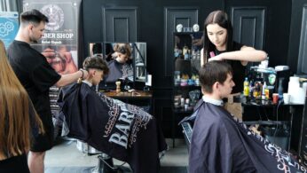 Dwóch młodych mężczyzn strzyże się w nowoczesnym salonie fryzjerskim. Fryzjer i fryzjerka, oboje ubrani na czarno, pracują nad swoimi klientami. W tle widoczne są czarne ściany, narzędzia fryzjerskie, lustra i markowe peleryny z napisem 