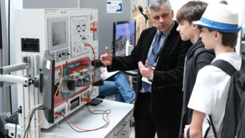 Mężczyzna w garniturze objaśnia techniczną maszynę z przewodami i ekranem dwóm nastoletnim chłopcom noszącym plecaki. Są w pomieszczeniu, prawdopodobnie na targach nauki lub imprezie edukacyjnej, z innymi ludźmi i sprzętem widocznymi w tle.