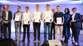 Grupa siedmiu młodych mężczyzn, w większości ubranych w białe koszule, stoi na środku sceny trzymając certyfikaty. Dorośli w formalnych strojach stoją po obu stronach, uśmiechając się. Światła sceniczne i fioletowe tło tworzą uroczystą atmosferę.