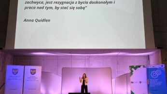 Kobieta stoi na scenie, prowadząc prezentację, a duży ekran za nią wyświetla cytat w języku polskim autorstwa Anny Quidlen. Scena jest otoczona dwoma pionowymi banerami z logo i tekstem, a delikatne fioletowe oświetlenie oświetla scenę.