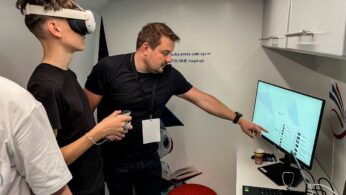 Osoba nosząca gogle VR trzyma kontroler, podczas gdy inna osoba obok niej wskazuje na monitor komputera wyświetlający listę. Znajdują się w nowoczesnym, białym pokoju z widoczną technologią i czerwonym stołkiem. Laptop i kubek znajdują się na biurku pod monitorem.