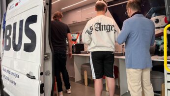 Trzech mężczyzn stoi wewnątrz nowoczesnej furgonetki przekształconej w miejsce do pracy. Drzwi furgonetki są otwarte, ukazując sprzęt i monitory w środku. Jeden z mężczyzn ma na sobie kurtkę z Los Angeles, a drugi niebieską marynarkę. Na zewnątrz furgonetki widoczny jest tekst oraz flagi UE i Polski.