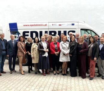 Grupa 23 dorosłych osób ubranych w stroje biznesowe stoi przed białą furgonetką z napisami "EDUBUS" i "www.opolskieLLL.pl". Pozują blisko siebie, uśmiechając się, z prostą białą ścianą w tle.