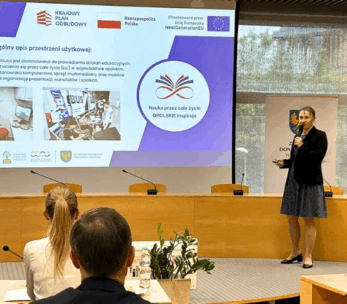 Kobieta w ciemnej sukience prezentuje pokaz slajdów w sali konferencyjnej. Slajd zawiera polski tekst, logo, zdjęcia pracujących ludzi i wspomina o Unii Europejskiej. Siedzący uczestnicy są zwróceni w jej stronę, a po prawej stronie wyświetlane są flagi Polski i UE.