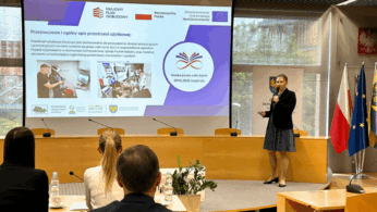 Kobieta stoi i przemawia na podium w sali konferencyjnej, prezentując slajdy na temat edukacji i szkoleń zawodowych. Cztery osoby siedzą naprzeciwko niej. Za nią znajdują się flagi Polski, UE i regionu oraz herb. Slajd prezentacji zawiera tekst, obrazy i logo.