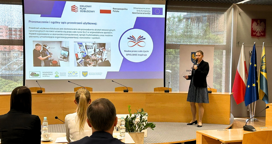 Kobieta stoi i przemawia na podium w sali konferencyjnej, prezentując slajdy na temat edukacji i szkoleń zawodowych. Cztery osoby siedzą naprzeciwko niej. Za nią znajdują się flagi Polski, UE i regionu oraz herb. Slajd prezentacji zawiera tekst, obrazy i logo.