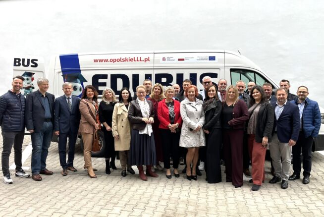 Grupa około 24 dorosłych osób, zarówno mężczyzn, jak i kobiet, pozuje uśmiechnięta przed białą furgonetką z napisem EDUBUS i www.opolskieLLL.pl. Stoją na zewnątrz na utwardzonej powierzchni, ubrani w stroje biznesowe i codzienne. Tłem jest zwykła biała ściana.