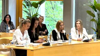 Cztery kobiety w strojach biznesowych siedzą przy drewnianym stole konferencyjnym z mikrofonami i dokumentami, skupione i zaangażowane. Za nimi siedzą ludzie, niektórzy z laptopami. Duże okna i zielone rośliny wypełniają jasną, nowoczesną salę konferencyjną.