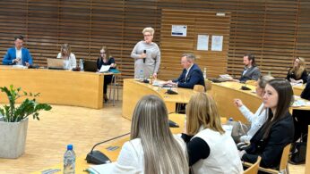 Kobieta stoi i przemawia w nowoczesnej sali konferencyjnej z drewnianymi ścianami. Stoi przed kilkoma siedzącymi osobami, zarówno mężczyznami, jak i kobietami, którzy mają przed sobą dokumenty, mikrofony i laptopy, co sugeruje formalną dyskusję lub prezentację.