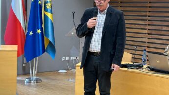 Starszy mężczyzna w czarnej marynarce i szarych spodniach mówi do mikrofonu w nowoczesnej sali konferencyjnej. Za nim znajdują się flagi Polski i UE oraz dwa herby na ścianie. Na drewnianym biurku po jego prawej stronie widoczny jest laptop.