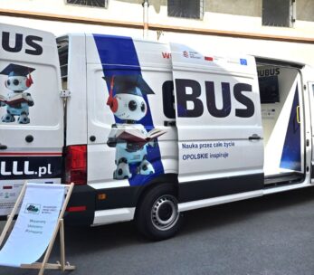Biała furgonetka z napisem EDUBUS z wizerunkiem robota w czapce absolwenta i trzymającego książkę. Boczne i tylne drzwi są otwarte, a z przodu stoi szyld z tekstem. Furgonetka jest zaparkowana w pobliżu budynku z zakratowanymi oknami.