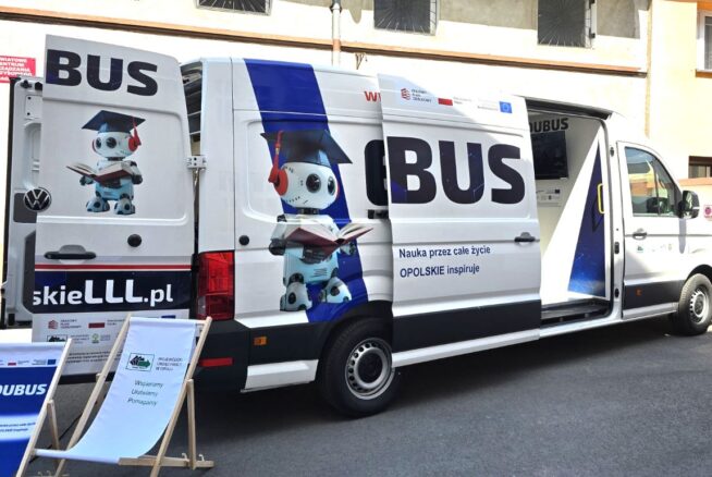 Biała furgonetka z napisem EDUBUS z wizerunkiem robota w czapce absolwenta i trzymającego książkę. Boczne i tylne drzwi są otwarte, a z przodu stoi szyld z tekstem. Furgonetka jest zaparkowana w pobliżu budynku z zakratowanymi oknami.
