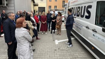 Grupa dorosłych stoi na zewnątrz w pobliżu białej furgonetki z napisem MOBUS. Grupa, ubrana w płaszcze i kurtki, uważnie słucha mężczyzny przemawiającego przy furgonetce. W tle widoczne są wysokie budynki z wieloma oknami.