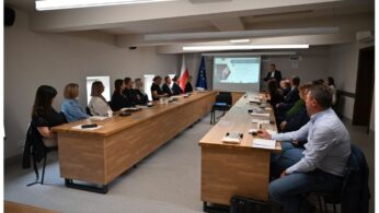 Grupa ludzi siedzi przy długich drewnianych stołach w sali konferencyjnej, uważnie obserwując prezentera z przodu przy ekranie projektora. W tle widać flagi, a ludzie robią notatki lub uważnie słuchają.