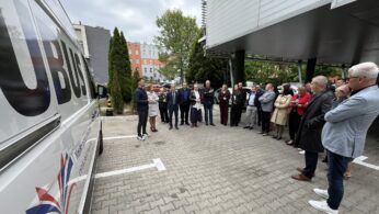 Grupa osób w strojach biznesowych stoi w półkolu na zewnątrz, uważnie słuchając mówcy w pobliżu białej furgonetki z częściowym napisem 
