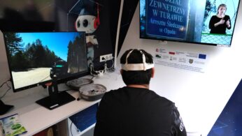 Osoba z czujnikami na głowie siedzi przy biurku naprzeciwko dwóch monitorów - jeden pokazuje leśną drogę z perspektywy pojazdu, drugi wyświetla polski tekst o ośrodku detencyjnym i tłumacza języka migowego. Na biurku znajduje się sprzęt elektroniczny i paczka chusteczek.