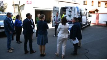 Grupa około ośmiu profesjonalistów stojących na zewnątrz w pobliżu białej furgonetki oznaczonej BUS z logo robota z kreskówki. Wygląda na to, że rozmawiają na parkingu obok budynku.