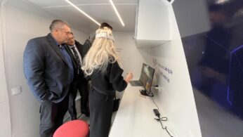 Trzej mężczyźni w garniturach obserwują kobietę z długimi blond włosami, noszącą urządzenie przypominające opaskę na głowie, wchodzącą w interakcję z monitorem dotykowym w nowoczesnym, jasnym pomieszczeniu z białymi ścianami i półkami. Na ladzie widoczne są urządzenia elektroniczne i kable.