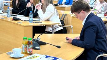Grupa ludzi siedzi uważnie w nowoczesnej sali wykładowej z drewnianymi krzesłami i biurkami. Niektórzy robią notatki lub przeglądają dokumenty, podczas gdy jedna osoba na pierwszym planie sprawdza swój telefon. Na biurkach leżą butelki z napojami, papiery i butelki z wodą.