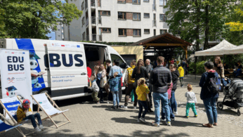 Grupa dorosłych i dzieci gromadzi się wokół białej furgonetki z napisem EDUBUS zaparkowanej na utwardzonym terenie w pobliżu budynku mieszkalnego. Niektóre dzieci noszą jaskrawe ubrania. W pobliżu ustawione są dwa składane krzesła z logo EDUBUS. Drzewa i namioty tworzą świąteczną atmosferę.