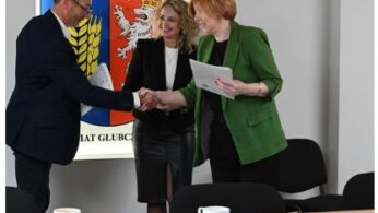 Trzy osoby w strojach biznesowych witają się w biurze. Dwie kobiety, jedna w zielonej marynarce, podają rękę mężczyźnie w ciemnym garniturze. Uśmiechają się, trzymając dokumenty. Za nimi na ścianie widnieje kolorowy emblemat przypominający herb. Na pierwszym planie znajduje się stół z filiżankami kawy.