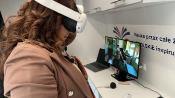 Kobieta z falującymi brązowymi włosami, ubrana w brązową marynarkę, korzysta z białego zestawu VR. Stoi przed biurkiem z monitorem komputera pokazującym osobę w podobnym zestawie słuchawkowym. Tło stanowią półki i polski tekst na ścianie.