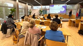 Grupa ludzi siedzi przy drewnianych stołach w nowoczesnej, jasnej sali konferencyjnej, słuchając kobiety prezentującej slajdy z przodu. Na stołach leżą filiżanki kawy, papiery i laptopy. W pomieszczeniu znajdują się duże okna, a w tle widoczne są flagi i rośliny.