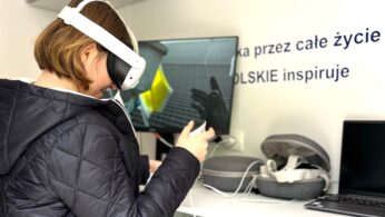 Osoba ubrana w czarną pikowaną kurtkę i biały zestaw VR trzyma kontrolery, siedząc przy biurku. Na biurku znajdują się akcesoria VR, laptop i monitor. W tle widoczny jest fragment napisu na ścianie w języku polskim.