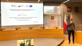 Mężczyzna w szarej marynarce stoi z mikrofonem w nowoczesnej sali konferencyjnej, zwracając się do publiczności. Za nim znajduje się duży ekran prezentacyjny wyświetlający slajd z polskim tekstem, logo i herbem. Widoczne są liczne flagi i podium.