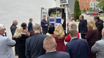 Grupa ludzi w strojach biznesowych stoi na zewnątrz w pobliżu białego budynku, słuchając mężczyzny wyjaśniającego przedmioty wewnątrz otwartej białej furgonetki. Furgonetka jest wypełniona pudełkami i materiałami eksploatacyjnymi. Większość uczestników jest odwrócona plecami do kamery i twarzą do mówcy.