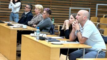 Pięć osób siedzi przy drewnianych biurkach w nowoczesnej sali wykładowej, uważnie obserwując coś poza kamerą. Obecne są trzy kobiety i dwóch mężczyzn; jedna kobieta w tle robi zdjęcia aparatem fotograficznym. Na biurkach znajdują się butelki z wodą, papiery i mikrofony.