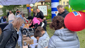 Ludzie gromadzą się wokół stołów na świeżym powietrzu przykrytych butelkami, torbami z prezentami i pudełkami pod baldachimem. Dorośli i dzieci przeglądają przedmioty i wchodzą z nimi w interakcje. Widoczne są kolorowe balony i plakaty informacyjne; w tle znajdują się budynki i zieleń.